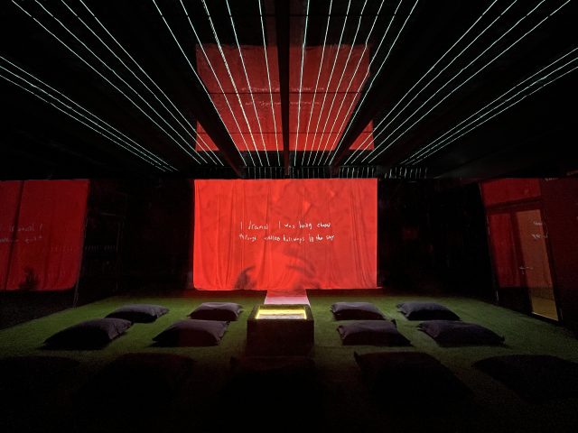 A projection screen is in the far end of a dark gallery. Astroturf and pillows are on the ground. The projection screen is on and shows white handwriting against a red background.