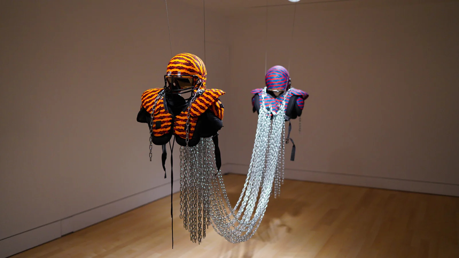 Two football helmets and pads suspended from the ceiling, facing away from each other, held together by chains.